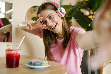 Close portrait of a young teen girl in headphones makes a selfie on the phone. cute Brunette sits in a cafe, listening to music with a phone and drinking juice.