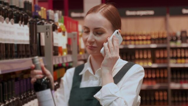 Young lady store customer talks on phone with friend. Red-haired woman chooses bottle of wine consulting with boyfriend on phone in department closeup