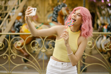 Charming girl playing with two colorful lollipops. Girl with pink hair posing with candies. carousel in the background. Rest in the amusement park