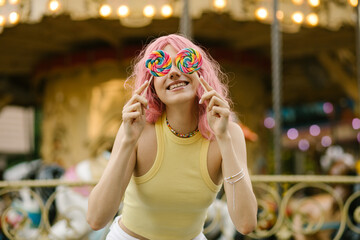 Charming girl playing with two colorful lollipops. Girl with pink hair posing with candies. carousel in the background. Rest in the amusement park
