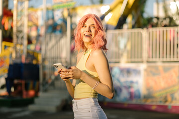 Young cute girl with pink hair posing on a carousel with a phone. Close portrait Charming hipster girl at sunset looks into the phone. Ferris wheel. Amusement park