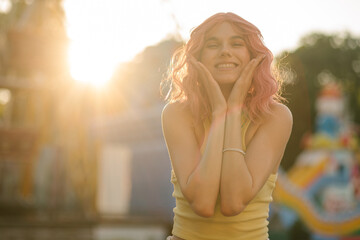 Young cute girl with pink hair posing on the background of the rides. Close portrait Charming hipster girl at sunset smiling