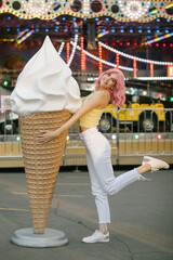 Cute Girl with pink hair posing with a big ice cream. Stylish girl in a yellow top. Attractions on the background
