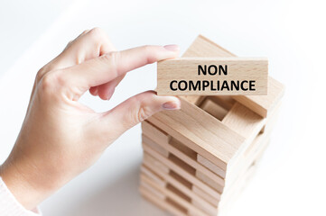 NON COMPLIANCE text on a wooden bar in a person's hand on a light background of a table