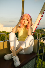 Close portrait of a girl listening to music in headphones. A happy girl with pink hair and a big smile. Amusement park in the background.