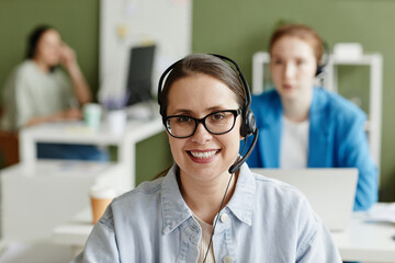 Portrait of young call centre agent in headphones and eyeglasses smiling at camera while working at...