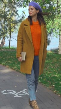 vertical video of happy woman in stylish autumnal outfit walking with book in park