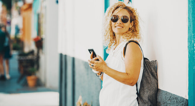 Happy female tourist pose for a picture using mobile phone connection against a white wall. Side portrait of woman smiling and writing on the cellular. Outdoor town leisure activity lady in summer
