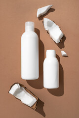 White cosmetic bottles, broken fresh coconut with sunny contrast shadows on brown background top view. Organic natural cosmetics with coconut oil. Plastic packaging products for branding, mock up