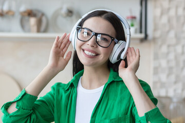 close-up photo portrait of young beautiful woman in glasses and green shirt smiling and looking at camera, brunette listening to music in big white headphones, resting at home