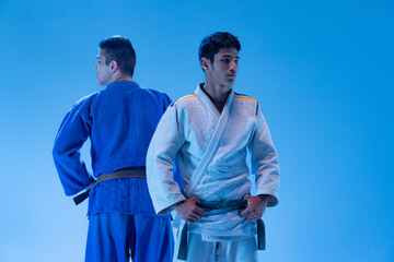 Portrait of two young sportive men posing in kimono isolated over blue studio background in neon light