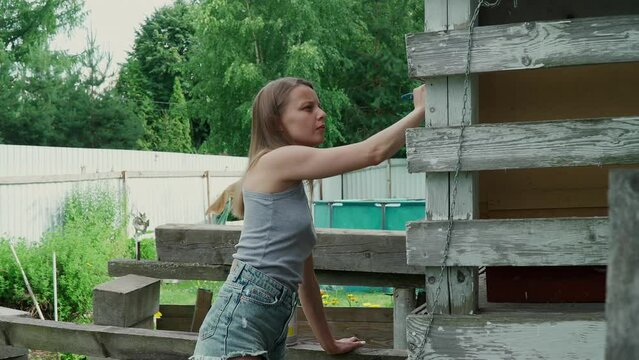 A beautiful caucasian girl in a gray tank top and short denim shorts paints the house with a brush. A European woman paints an old porch with white paint. painting work in a country house.