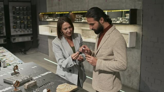 Tracking in of female Caucasian salesperson consulting young bearded Biracial man by display in luxury jewelry shop, choosing precious ring
