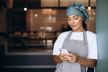 Woman baker in apron standing with phone by the doors at the coffee shop