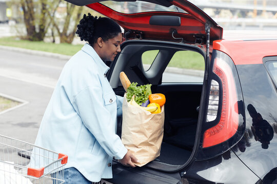 Side view portrait of smiling black woman putting groceries in car trunk in parking lot, copy space