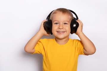 A cute little smiling boy in a yellow T-shirt on a light gray background with black headphones enjoys listening to music.