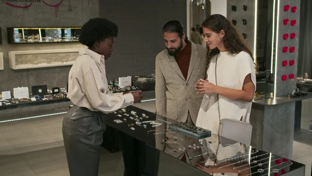 Medium long of young Biracial hetero couple shopping for ring in modern jewelry store, Black female salesperson helping