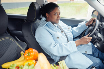 Portrait of adult black woman driving car with grocery bag in foreground after shopping in...
