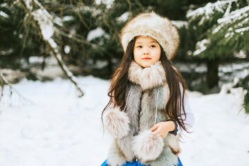 portrait of beautiful cute little Asian girl in fur coat and fur hat standing in snowy forest in...