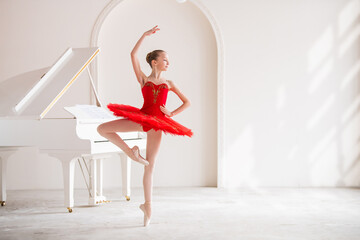 A cute little girl dreams of becoming a professional ballerina. In a white room, next to a piano, a girl in a bright red tutu is dancing on pointe shoes. Vocational school student.