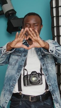 Vertical video: African american photographer doing heart shape symbol with hands, psoing with romantic love gesture over background with photography lighting equipment. Valentines day feelings sign.