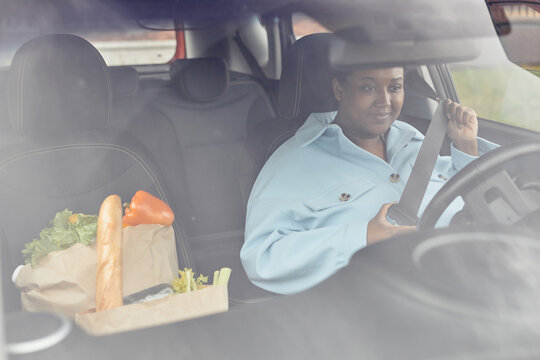 Behind Glass View Of Black Woman Putting On Seatbelt In Car After Shopping For Groceries, Copy Space