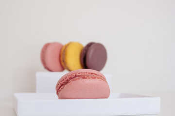Close-up of pink macaron in foreground and mix of appetizing blurred macarons on white box in background. Light background. 