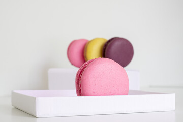 Close-up of pink macaron in foreground and mix of appetizing blurred macarons on white box in background. Light background. 