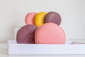 Close-up of appetizing pink and brown macaron in foreground and mix of blurred macarons on white box in background. Light background. 