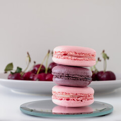 Close-up of appetizing mix of macarons with blurred plate with red cherries in background on white background.