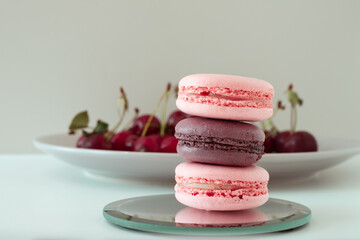 Close-up of appetizing mix of macarons with blurred plate with red cherries in background on white background.