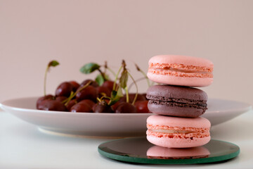 Close-up of appetizing mix of macarons with blurred plate with red cherries in background on white background.