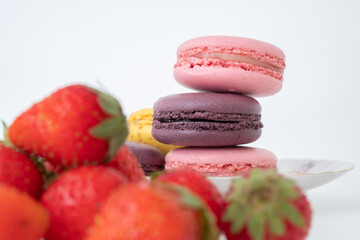 Mix of appetizing macarons on plate on white background with fresh strawberries in foreground 