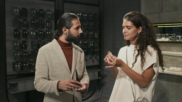 Medium of young curly-haired woman and bearded Biracial man standing by display of designer jewelry in luxury shop, picking earrings, talking