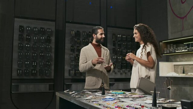 Medium long of young curly-haired woman and bearded Biracial man standing by display of designer jewelry in luxury shop, talking