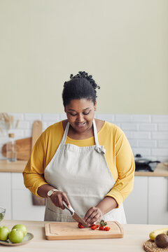 Vertical portrait of smiling black woman cooking healthy meal in kitchen and cutting vegetables