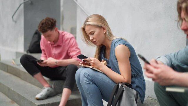 Group of teenagers sitting in outdoor stairs in front of school. Everybody using phone, scrolling. Internet and social media addiction among teenagers concept.
