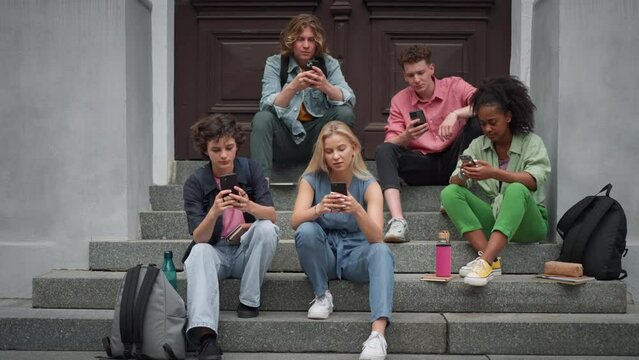 Group of teenagers sitting in outdoor stairs in front of school. Everybody using phone, scrolling. Internet and social media addiction among teenagers concept.