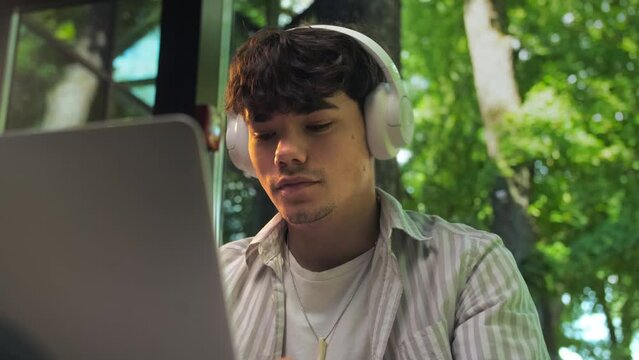 Serious Young man puts on headphones and working at his computer in a restaurant against a backdrop of green trees. man smiles and listens to music sitting in a cafe.