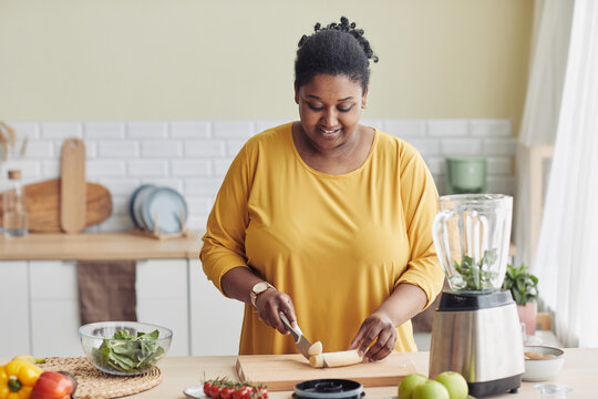 Portrait of smiling black woman cutting bananas while making healthy meal in kitchen and filming cooking video, copy space