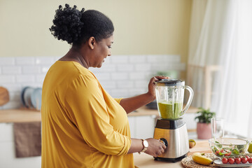 Side view portrait of overweight black woman using blender while making healthy smoothie at home kitchen, copy space