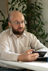 Millennial man with glasses in light shirt using mobile phone in his hand, working from home