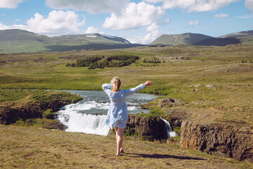 Woman wearing blue summer dress in wild nature in Iceland, Europe. Green mountains and waterfall on background, blue sky, sunny day.