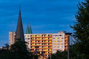 Tall block of flats from the 20th century with 11 floors and orange balcony parapets. Windows and facade reflecting orange sunset sky. Towers of churches in town centre of Iserlohn Sauerland Germany.