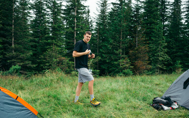 Young male tourist is standing on a mountain near a campsite with tents on a forest background and looking at the camera