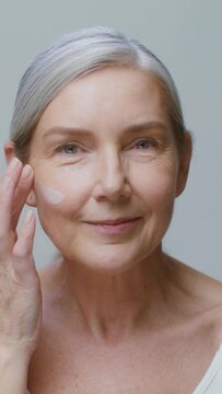 An older model applies moisturizer to her face while smiling at the camera