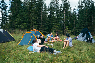 Group of people are resting in the mountains with tents. Photo of a group of tourists friends in a campsite with tents on the background of the forest are sitting and talking.