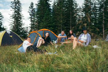 Group of young people tourists near a campsite with tents in the mountains sitting on the grass and looking at the camera and posing.