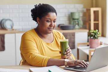Portrait of overweight black woman drinking smoothie and using laptop at home - obrazy, fototapety, plakaty