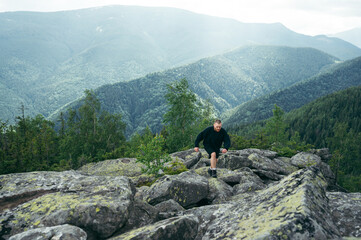 Focus on the landscape. Active young man in casual clothes is climbing a rock in the mountains at the top and looking at the camera.